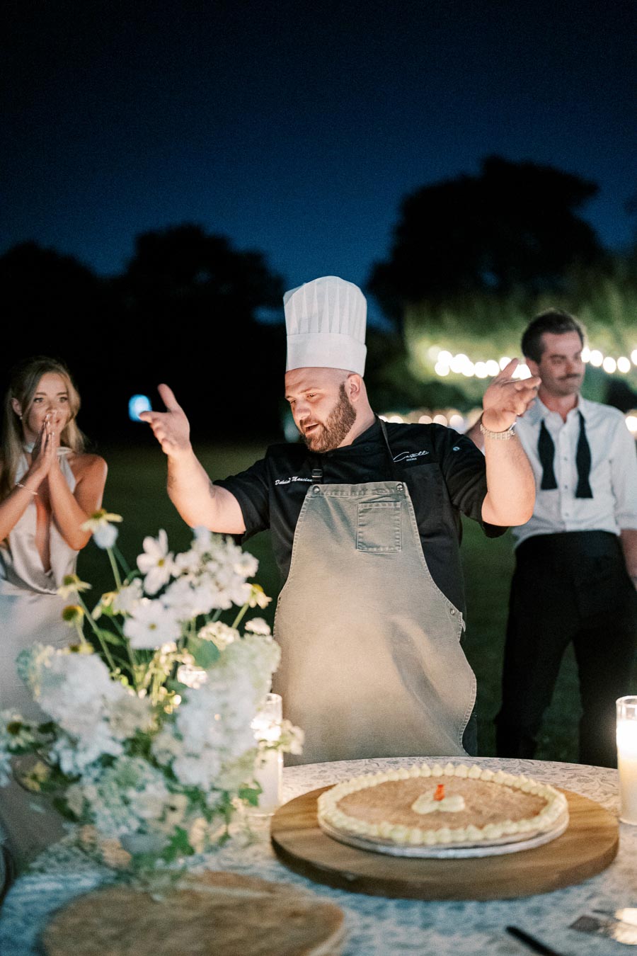 Chef celebrating outdoors with guests, presenting a decorated cake on a table adorned with flowers and candles, against a serene evening backdrop.