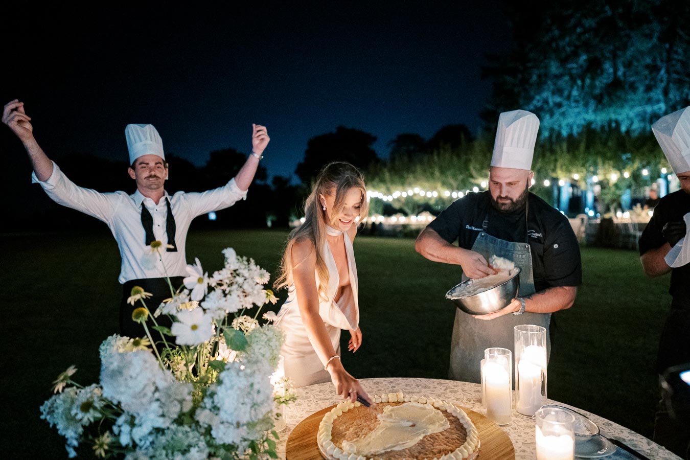 A joyful wedding reception at night featuring a bride spreading frosting on a large cake, assisted by chefs in hats. Romantic candlelight and floral decorations enhance the festive outdoor setting.