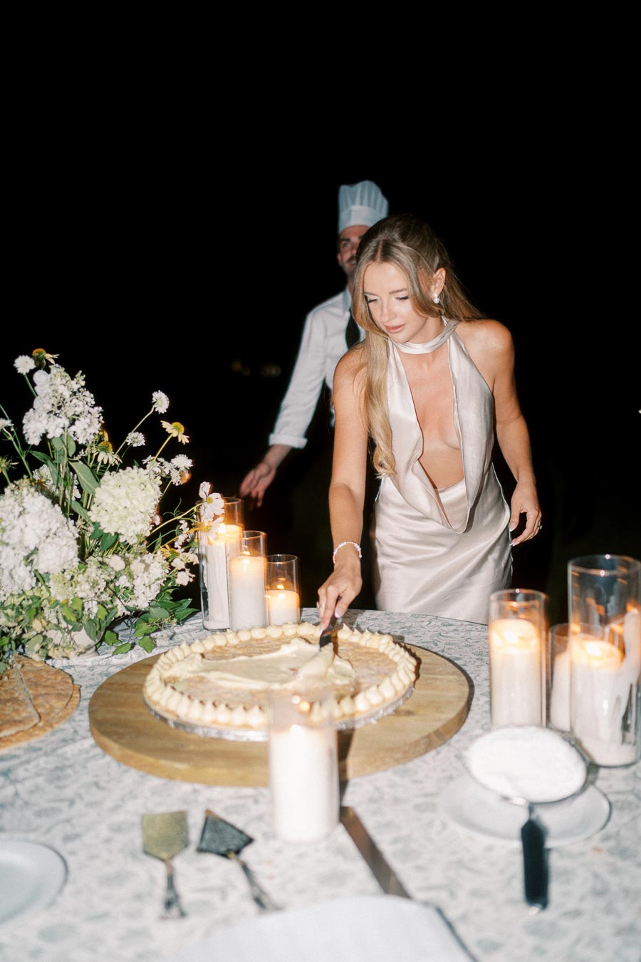 A woman in an elegant dress slices a large cake on a beautifully decorated table with flowers and candles, creating a romantic ambiance at a nighttime event.
