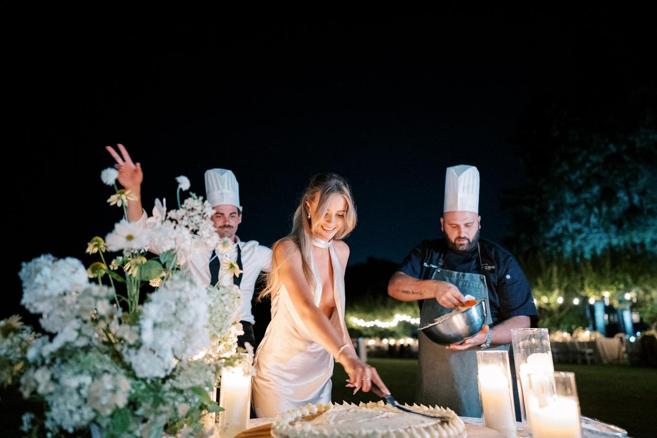 A bride in a white dress cuts a wedding cake outdoors at night, surrounded by chefs in white hats, with decorative candles and flowers in the foreground, set against a backdrop of string lights.