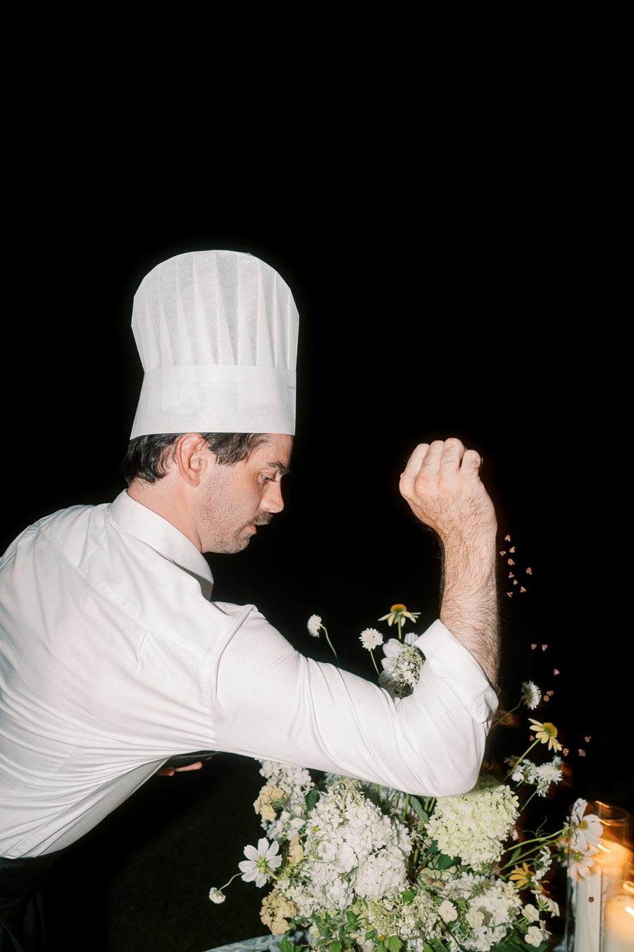 Chef in a white uniform and tall hat stylishly sprinkling seasoning, with a dark background and a vibrant display of fresh flowers.