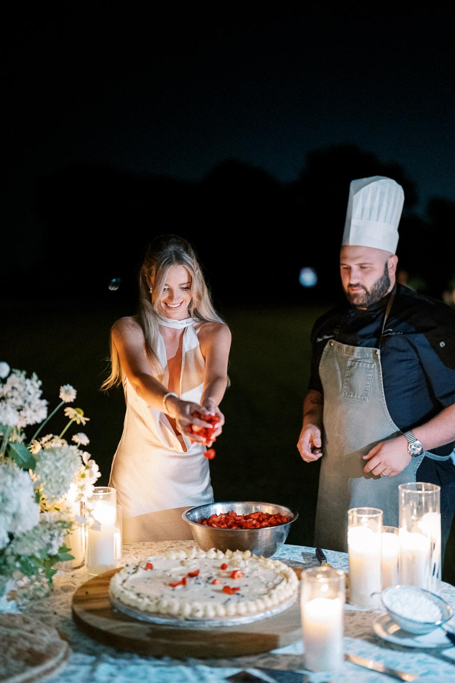 A woman in a white dress adds strawberries to a large cake outdoors at night, assisted by a chef in a tall hat. Candles and flowers decorate the table, creating a warm and festive atmosphere.