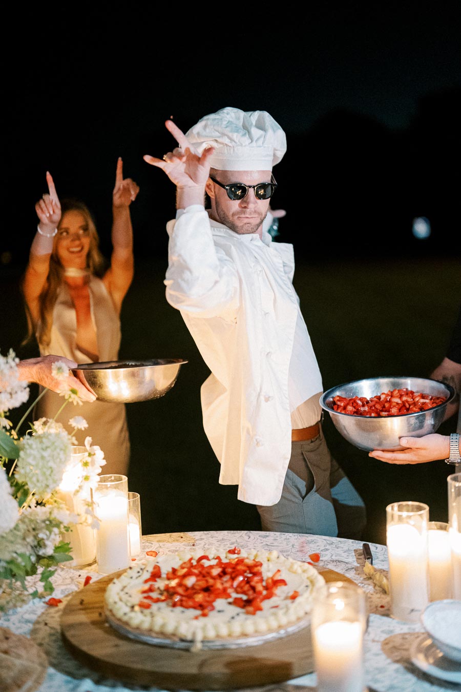 Chef in a white uniform and sunglasses theatrically posing while holding a bowl of strawberries, with a festive atmosphere including lit candles, a dessert table, and guests celebrating in the background.