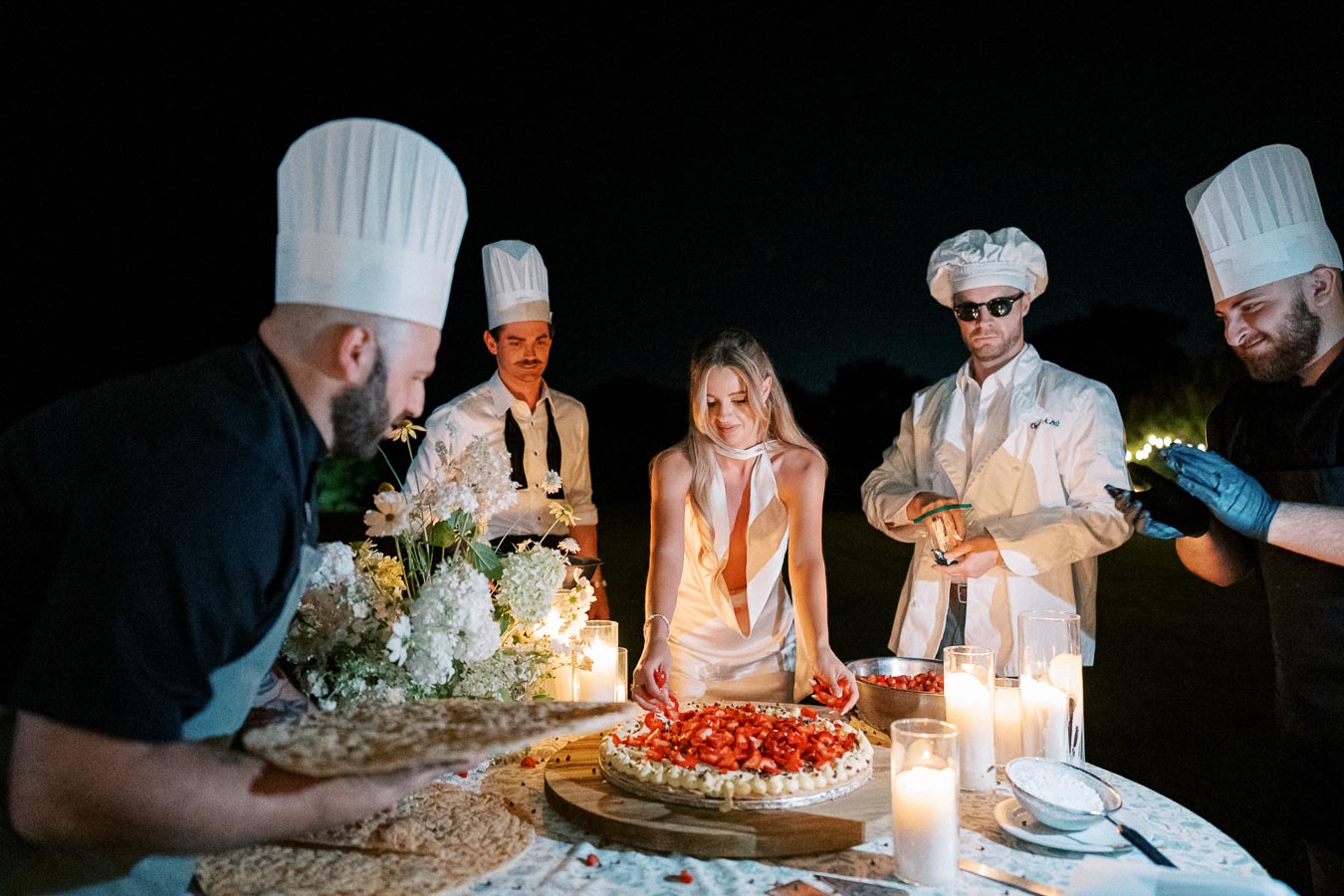 A group of chefs and a woman gather around a beautifully decorated cake, topped with strawberries, at an elegant outdoor event at night. Candles and white flowers adorn the table, adding to the festive ambiance.