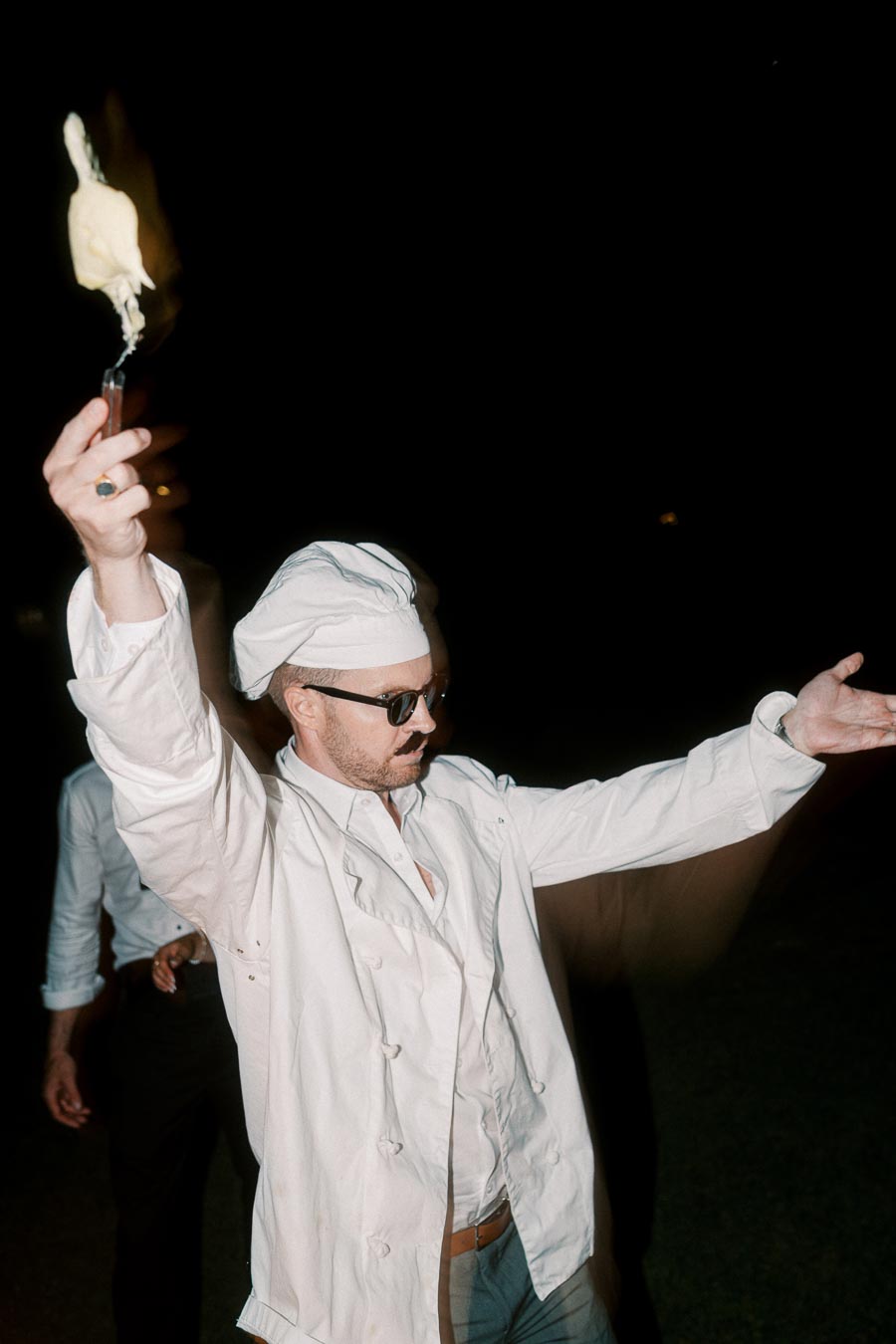 A chef wearing sunglasses and a white uniform holds a flaming torch in one hand, standing confidently against a dark background.