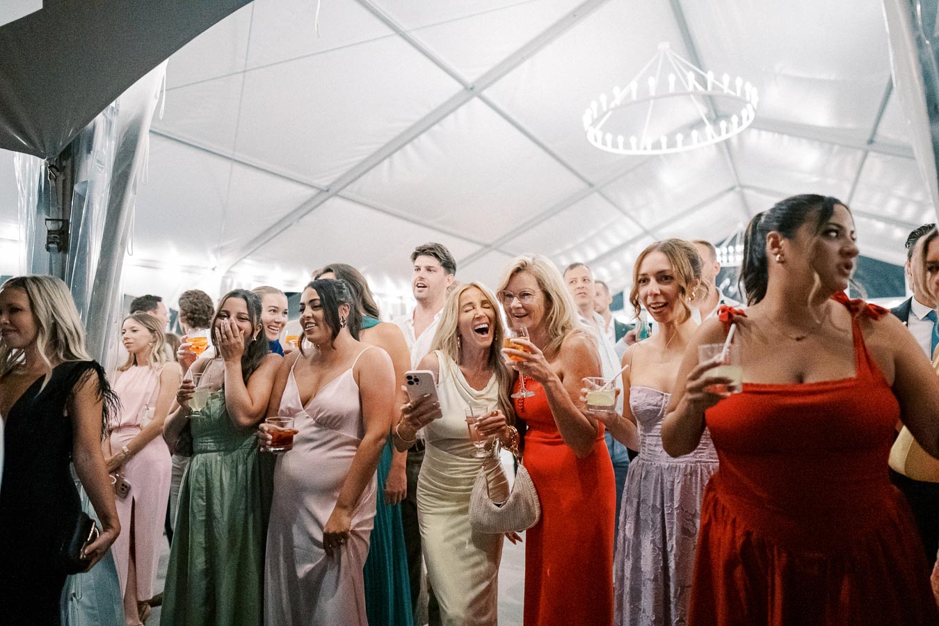 A group of elegantly dressed people enjoying a lively gathering in a large tent, holding drinks and smiling, with a chandelier visible in the background.