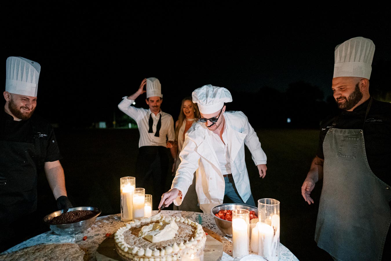 Chefs wearing hats and aprons collaborate at a nighttime outdoor event, decorating a cake surrounded by candles and bowls of ingredients.