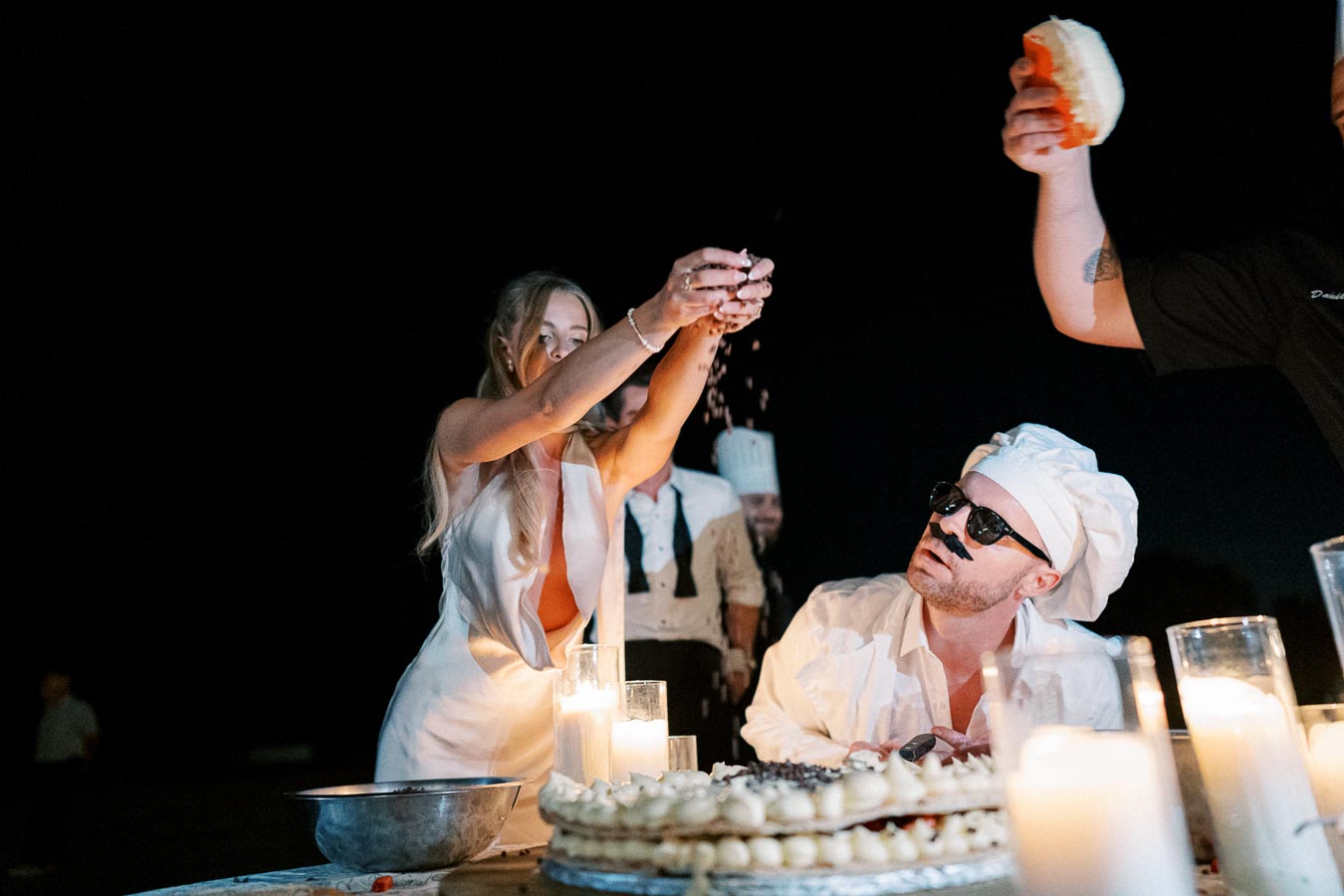 A lively nighttime outdoor scene featuring a woman in a white dress sprinkling something over a large cake, with a man wearing a chef’s hat and sunglasses sitting nearby. Candles illuminate the table, creating a warm atmosphere.