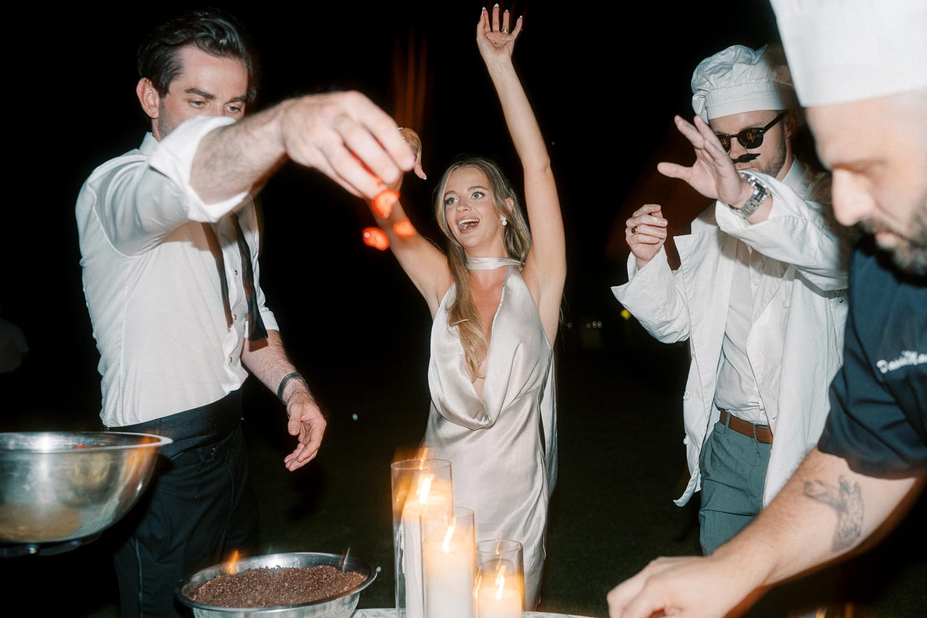 A group of people dressed in formal and themed attire celebrate at an outdoor event. A woman in a satin dress has her arms raised, surrounded by festive lighting and decorative candles, as other attendees participate in the joyful atmosphere.