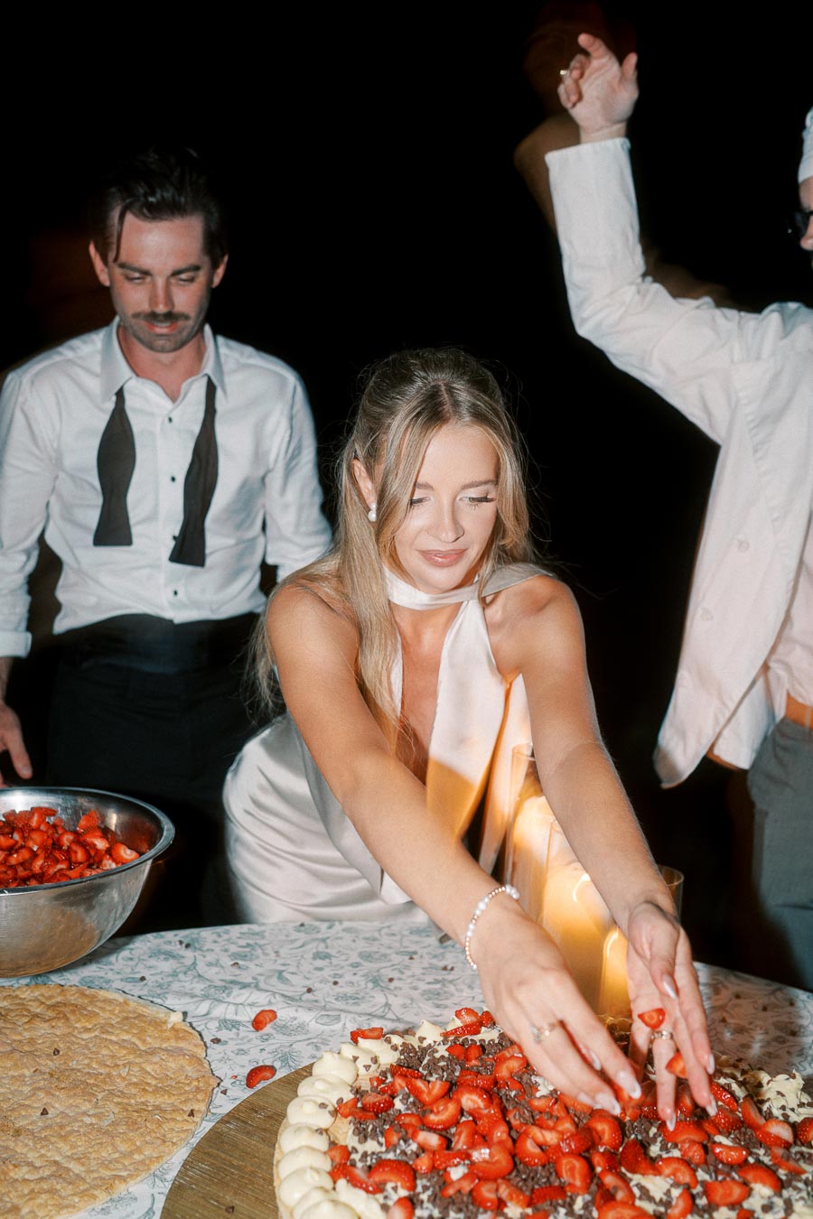Woman in a white satin dress preparing a large dessert with strawberries and cream at a festive gathering, accompanied by friends in formal attire with candlelight ambiance.