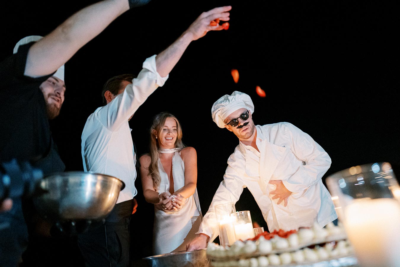 Group of people joyfully engaged in an outdoor culinary experience at night, with a chef in glasses and a white hat tossing food into the air, creating a lively and festive atmosphere.