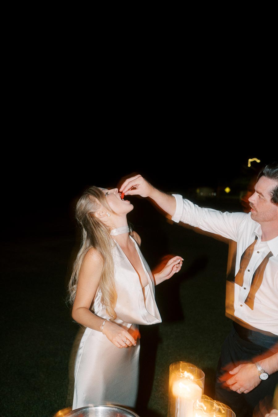 A woman in an elegant white dress eats a strawberry fed by a man in a black and white tuxedo, with candles setting a warm ambiance at night.