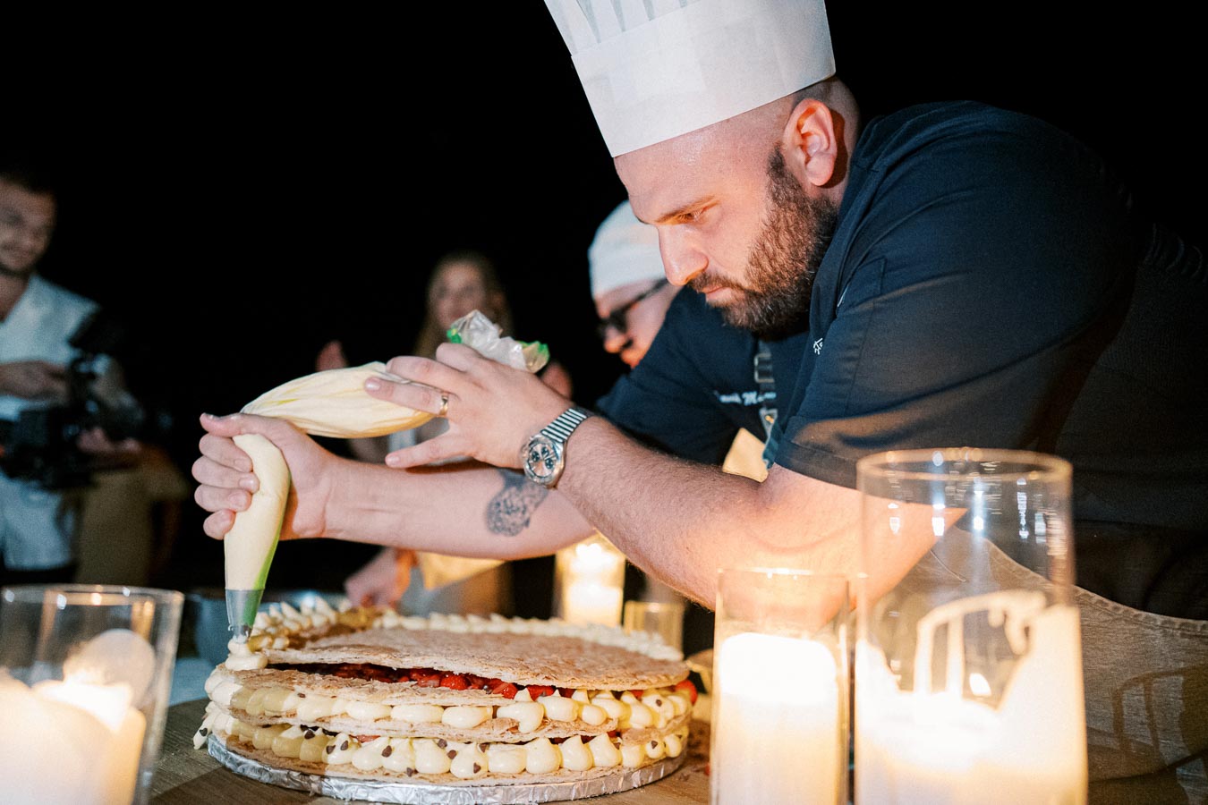 Pastry chef meticulously decorating a large layered cake with cream using a piping bag, surrounded by glowing candles for ambiance.