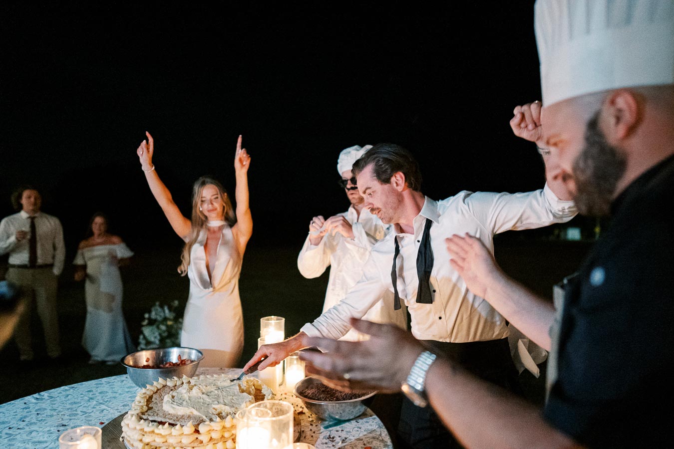Wedding celebration at night with bride and guests dancing joyfully around a cake table, decorated with lit candles, as a man in formal wear cuts the cake.