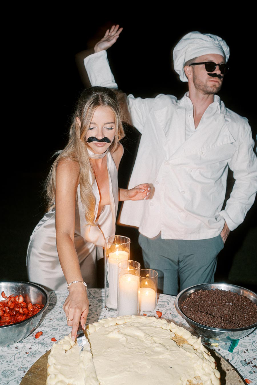 A woman in a satin dress and fake mustache cuts a large cake at an outdoor nighttime event, with a man dressed as a chef standing beside her. The table is decorated with candles and bowls of strawberries and chocolate chips.