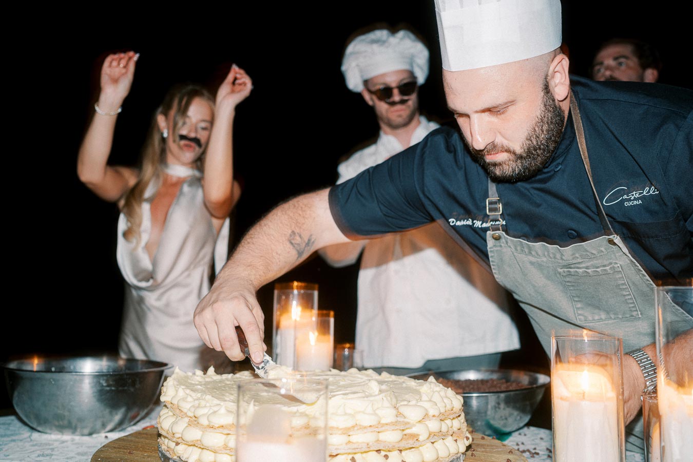 Chef in a white hat and apron decorates a multi-layered cake, surrounded by candlelight, while people in festive attire celebrate in the background.