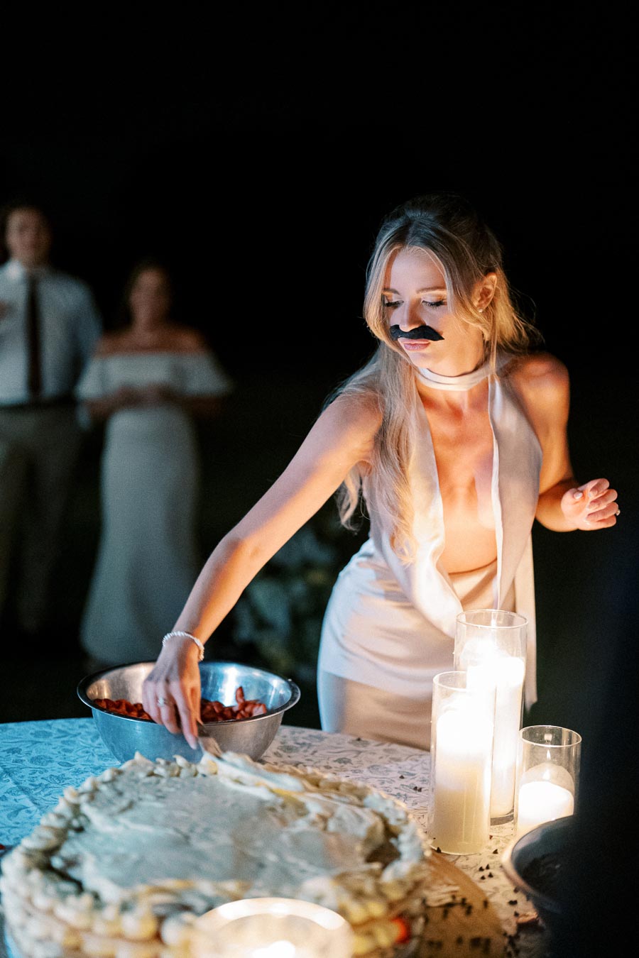 A woman with a fake mustache in a white dress serves cake at a candlelit event, with guests watching in the background.