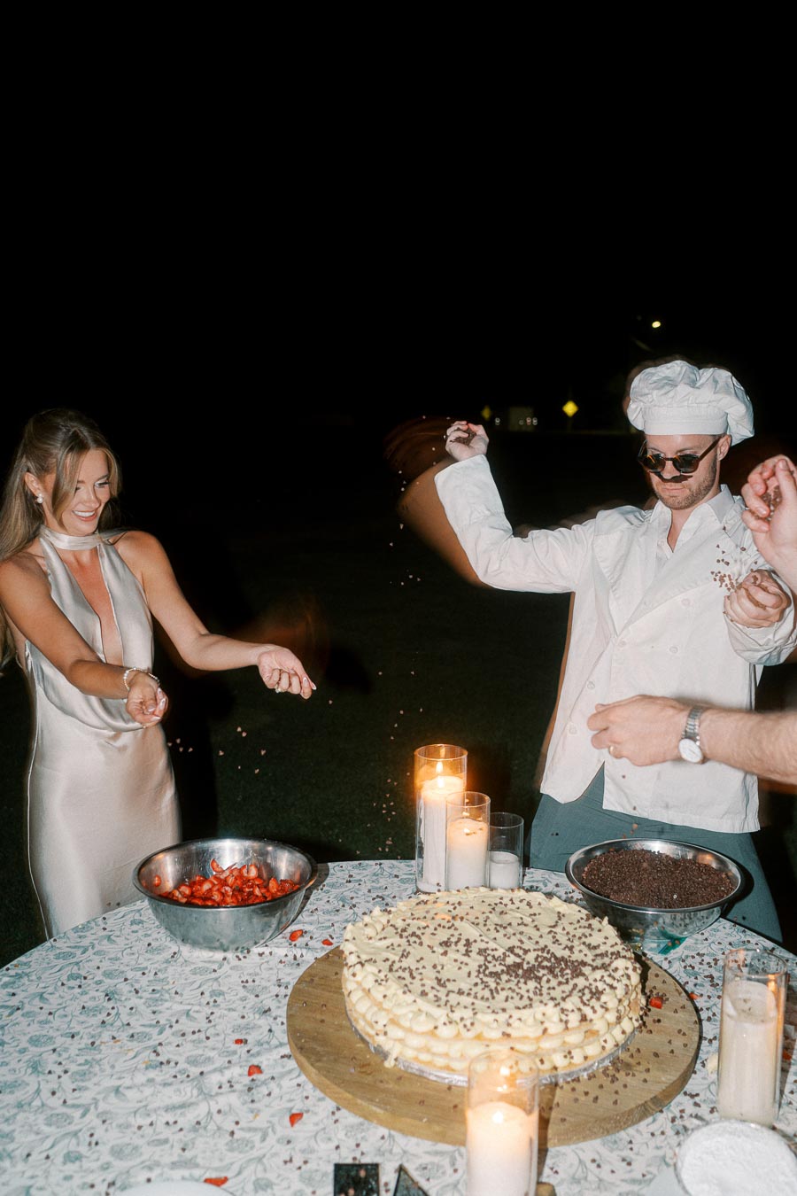 A woman in an elegant dress and a man dressed as a chef are joyfully sprinkling ingredients onto a large cake on an outdoor table at night, with candles illuminating the scene.