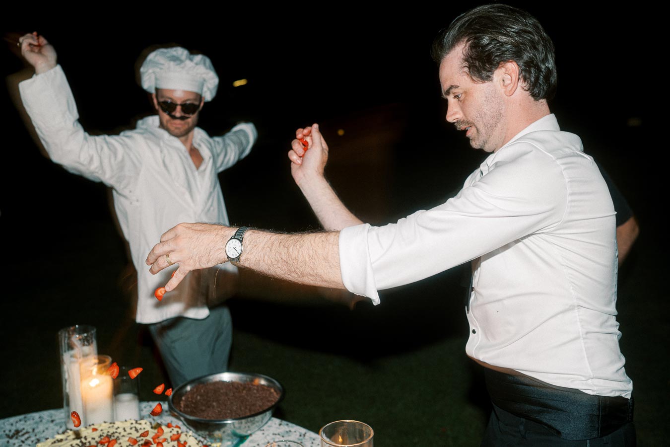 Two men enthusiastically decorating a cake at night, one wearing a chef's outfit and sunglasses, with strawberries being sprinkled on the dessert.