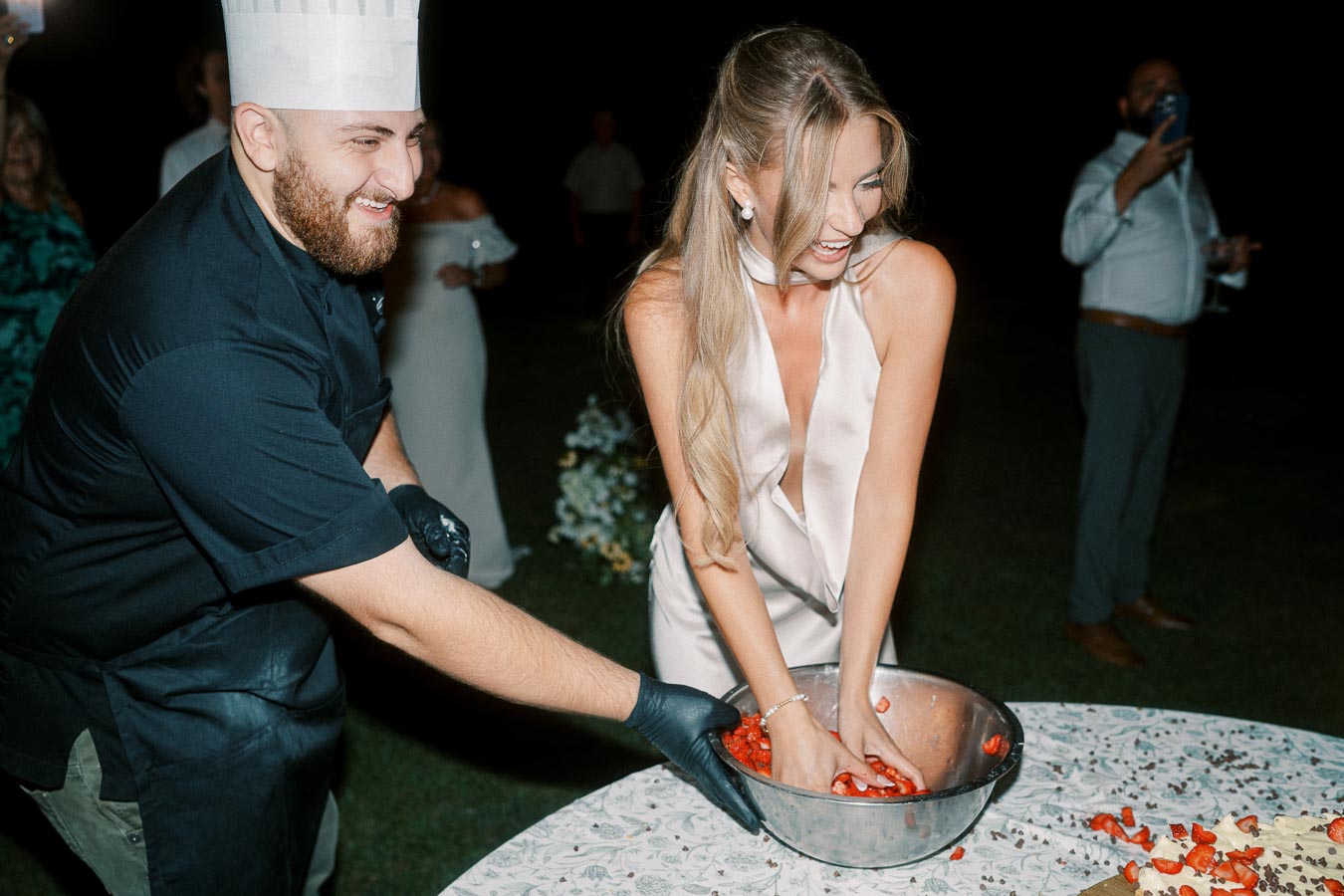 A joyful woman in a white dress and a smiling chef in a hat are playfully mixing strawberries in a metal bowl at an outdoor evening event, surrounded by onlookers and a decorated table.