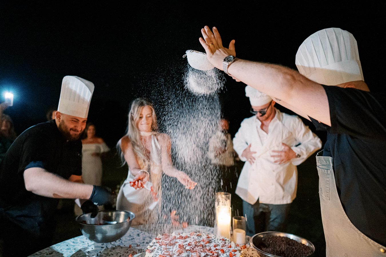A group of chefs and a woman joyfully preparing dessert outdoors at night, with flour being dramatically sprinkled over a table filled with ingredients and lit candles, creating a festive culinary scene.