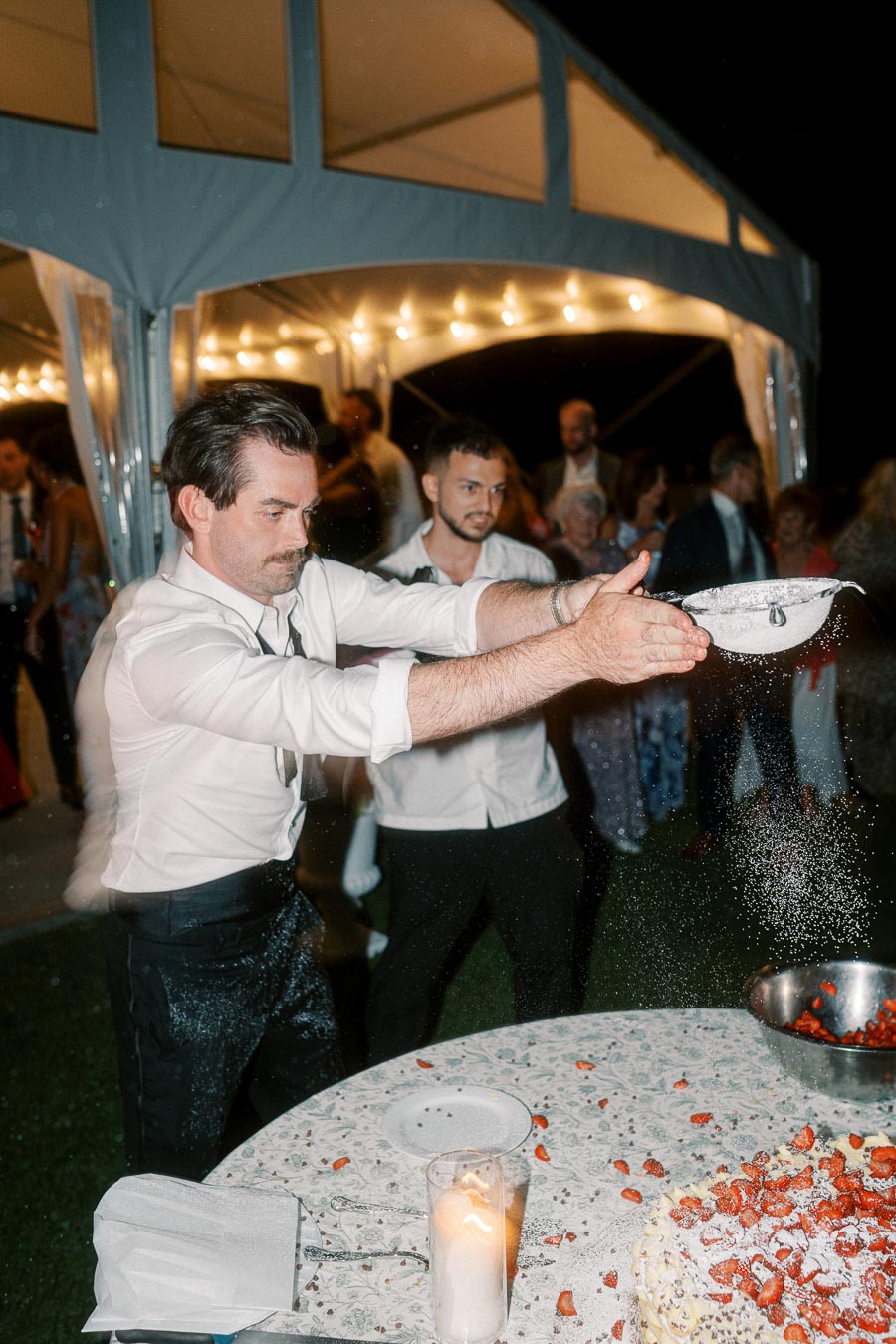 Man in white shirt enthusiastically sifting powdered sugar onto a cake at an outdoor event, with a group of people mingling under a lit tent in the background.