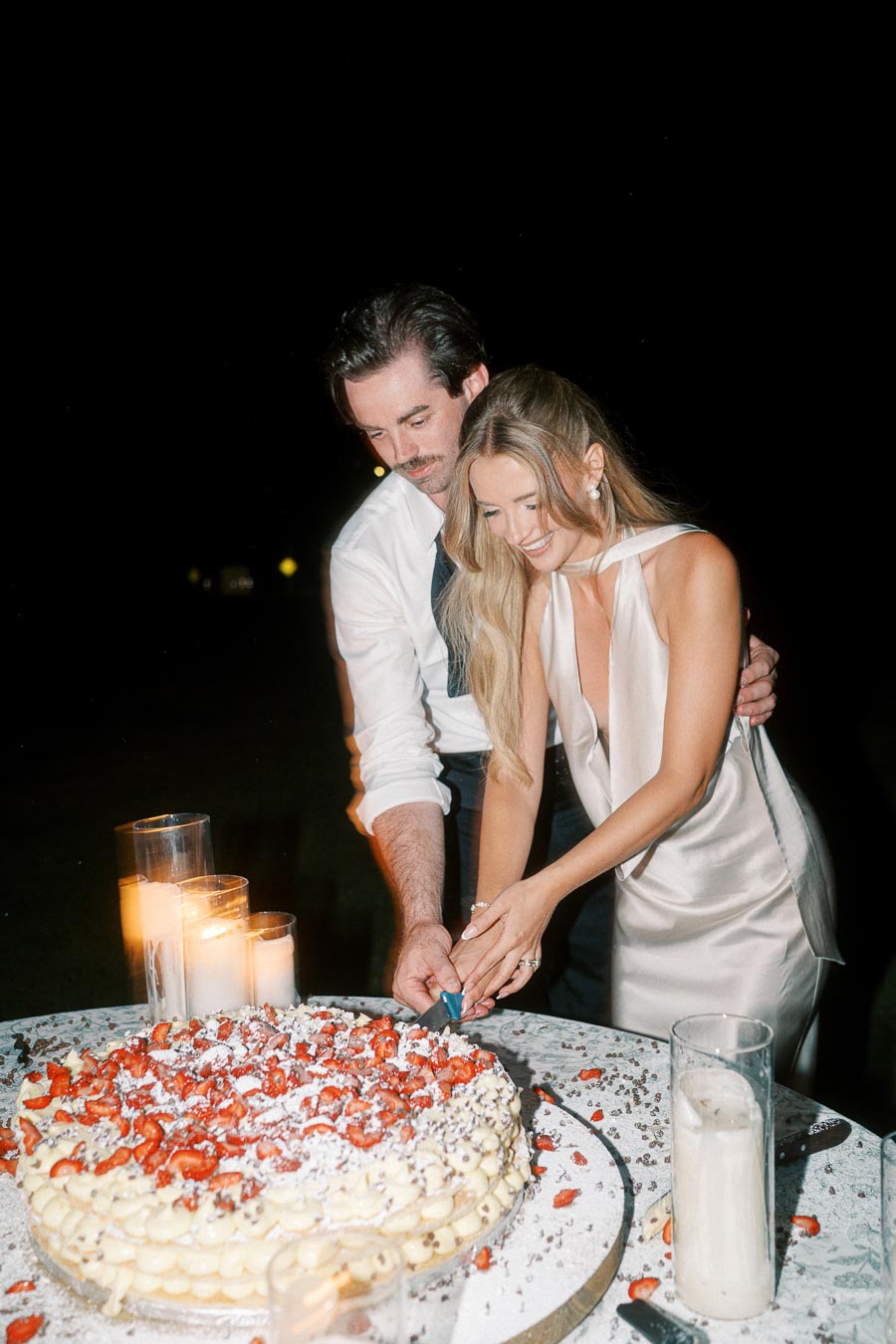 Couple cutting a large celebratory cake topped with strawberries and cream at night, surrounded by lit candles on a table.
