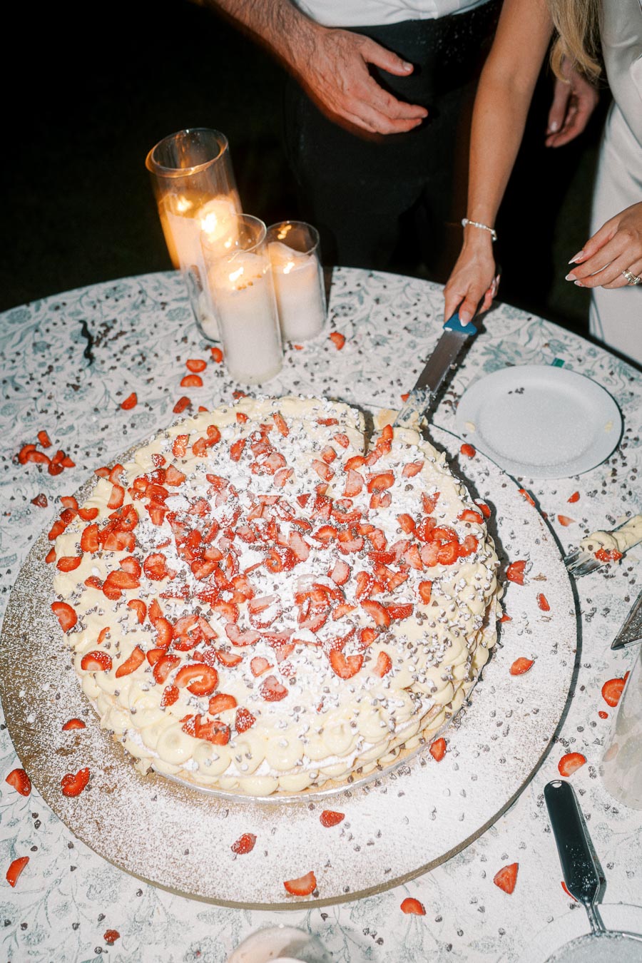 A beautifully decorated strawberry-topped cake on a floral-patterned tablecloth, with hands cutting into it surrounded by glowing candles, creating a warm, festive atmosphere.