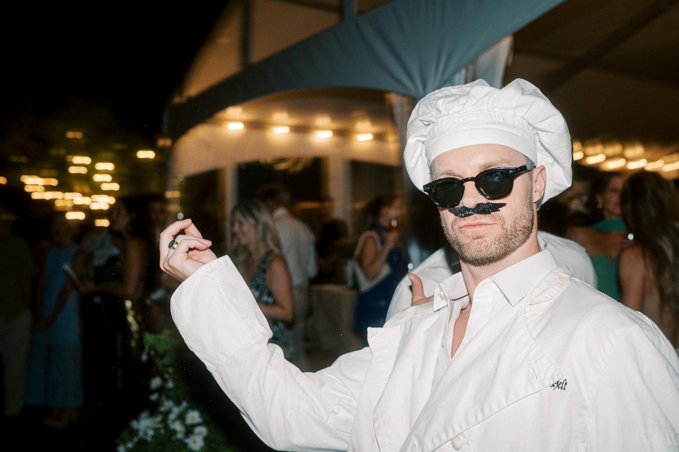 Man in a white chef's outfit and sunglasses striking a playful pose at a lively night event.