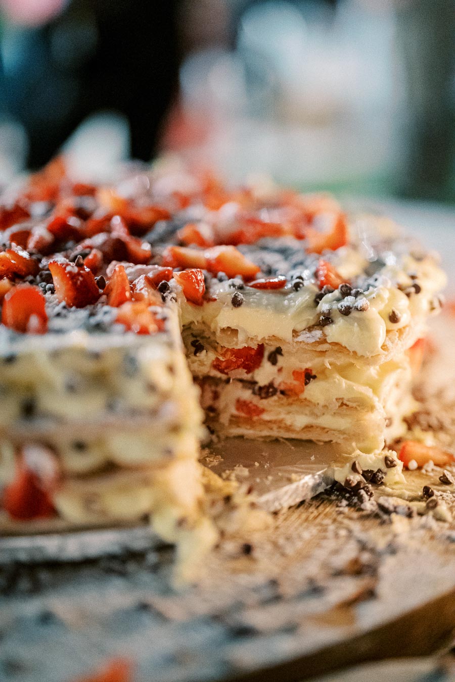 A close-up of a delicious layered cake filled with fresh strawberries and cream, topped with chocolate chips and strawberry pieces, on a wooden table.