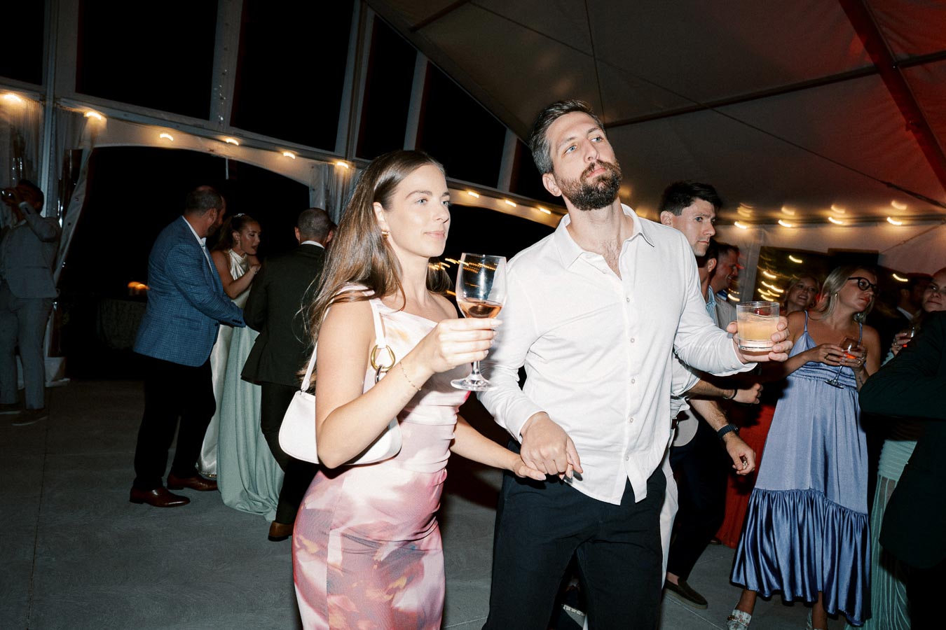A group of people dancing and enjoying drinks at an evening party under a large, decorated tent. The atmosphere is lively with warm lighting and elegant attire.