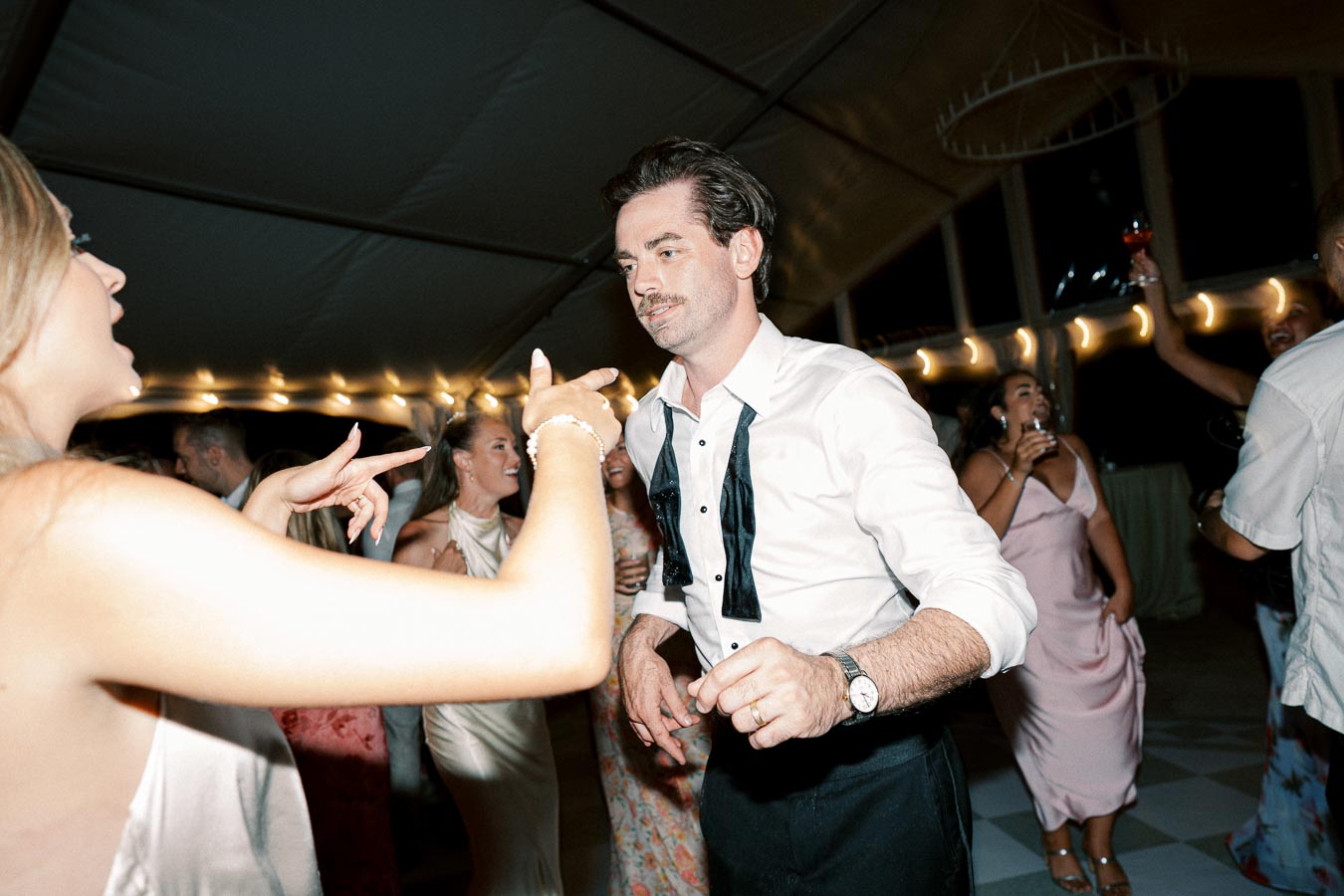 Group of people dancing energetically at a lively event, with a man in a white shirt and loosened black tie at the center. The atmosphere is festive, with soft lighting and guests enjoying the celebration.