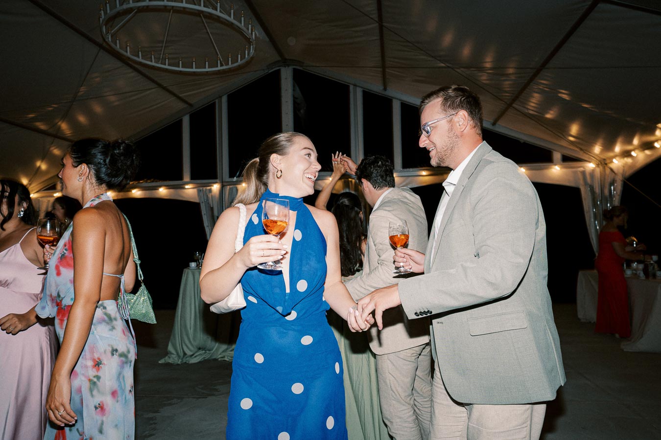 A group of people enjoying a festive evening party under a large tent with string lights, holding drinks and engaging in conversation.