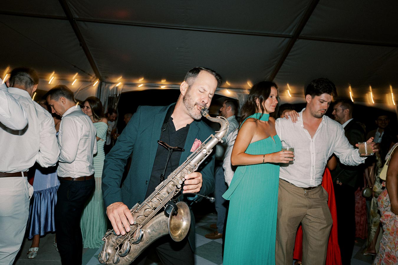 A lively wedding reception with a saxophonist performing energetically, surrounded by elegantly dressed guests dancing under a beautifully lit tent.
