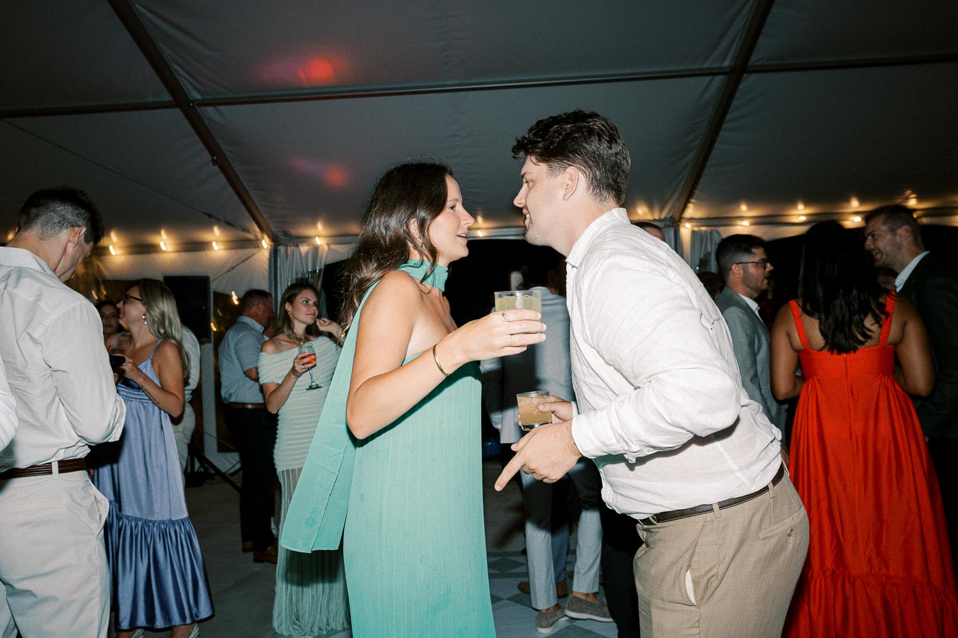 Couple enjoying a lively party in a tent with friends, holding drinks and engaged in cheerful conversation, surrounded by elegantly dressed guests.