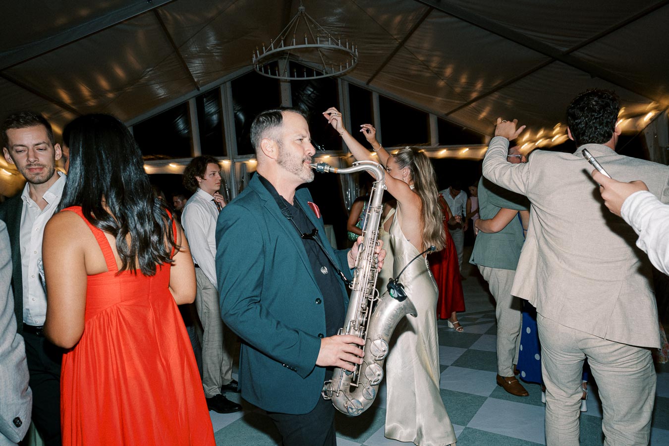 A saxophonist playing lively music at a wedding reception, surrounded by elegantly dressed guests dancing under a canopy with string lights.