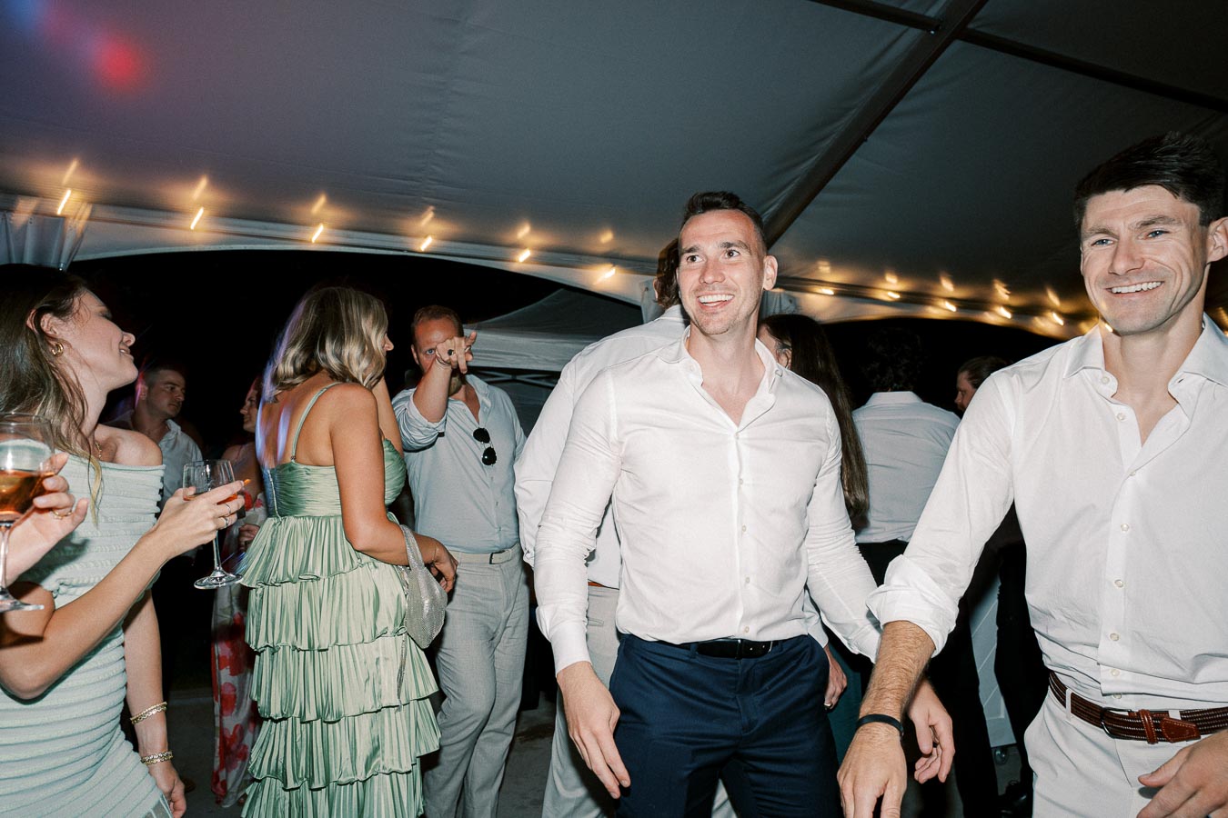 A group of well-dressed people enjoying a lively party under a tent with string lights, smiling and holding drinks.