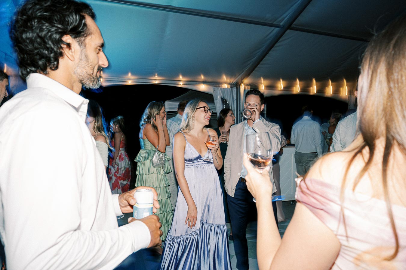 People enjoying a lively outdoor evening event under a decorated tent, holding drinks and socializing in elegant attire.