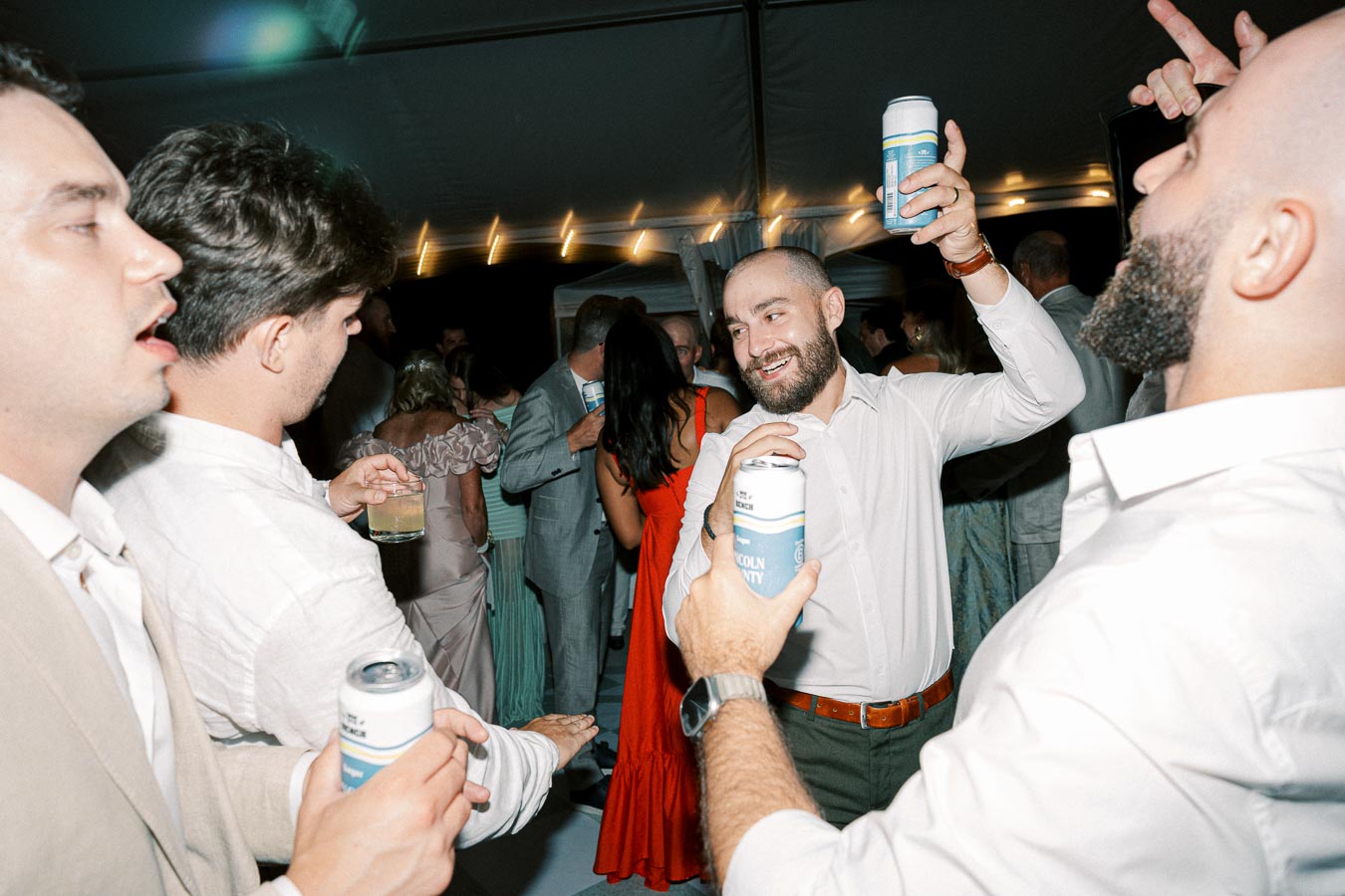 Group of people enjoying a lively party, holding drinks and engaging in conversation under a tent with string lights.