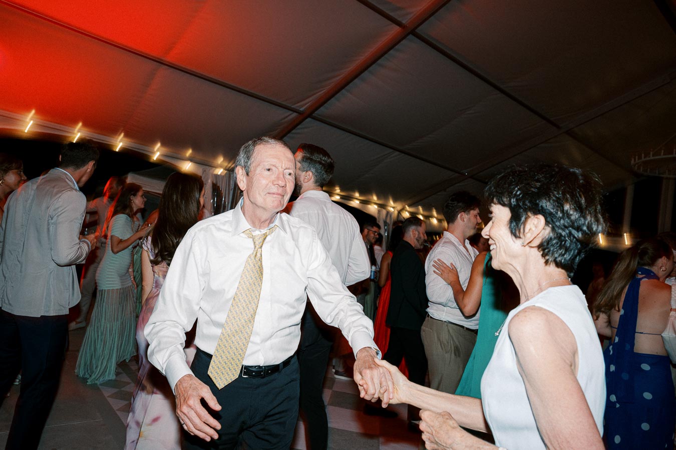 Senior couple joyfully dancing at a lively evening event under a tent with colorful lights and a cheerful crowd in the background.