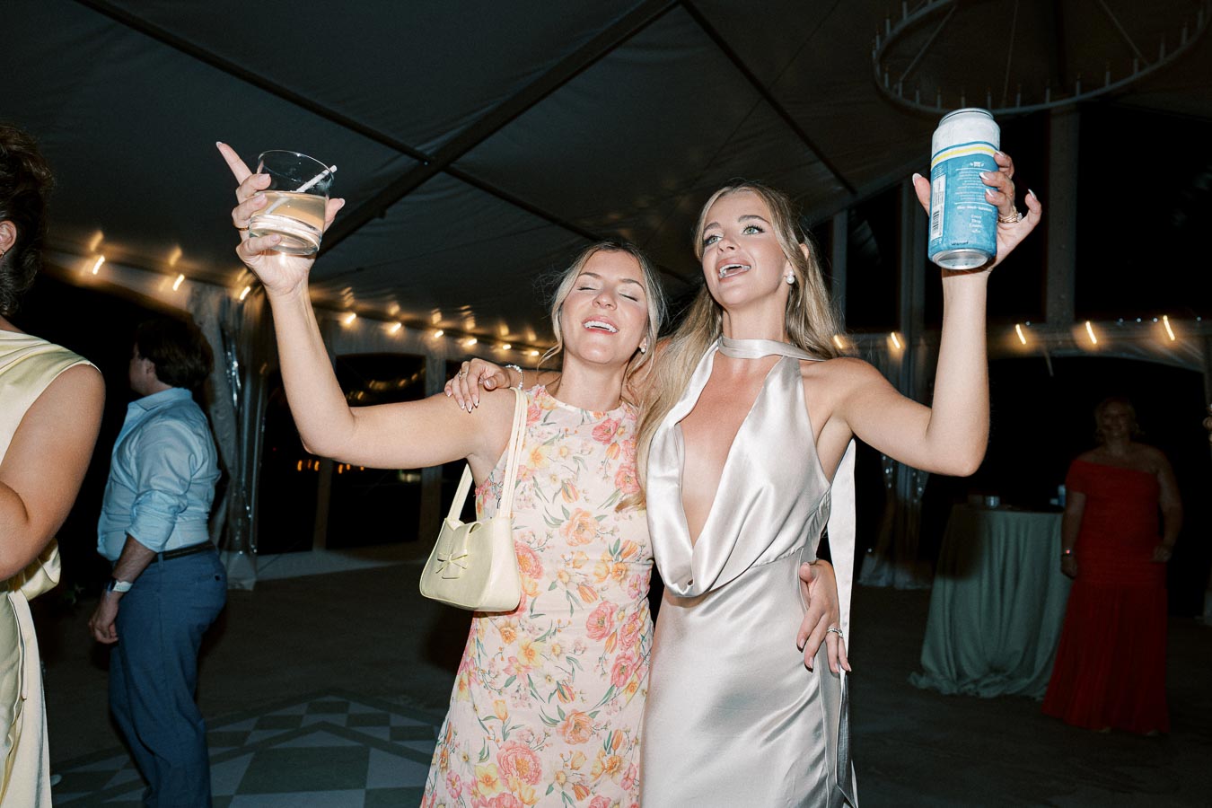 Two women enjoying a lively celebration under a lit tent, holding drinks and dressed elegantly for the occasion.