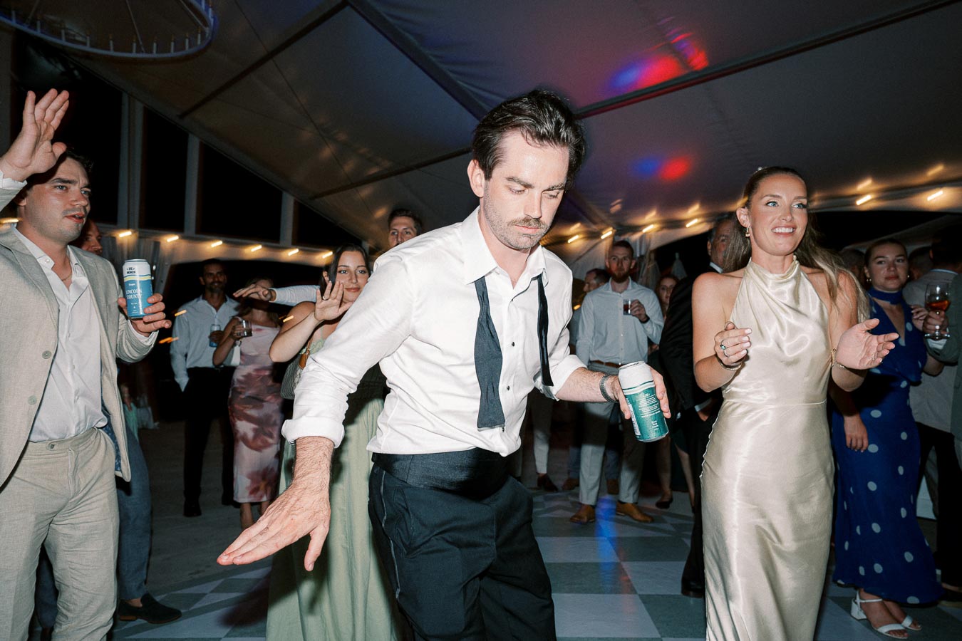 Group of people dancing energetically under a marquee tent with soft lighting at a lively party, featuring a man in a loosened tie and rolled-up sleeves, holding a canned drink.