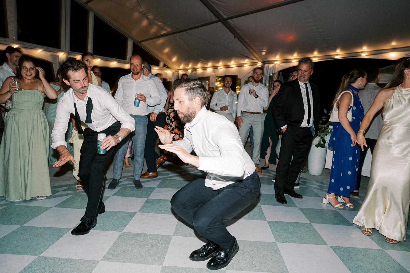 A lively wedding reception dance floor scene under a tent with guests dressed in formal attire, showcasing two men energetically dancing in the center while others watch and enjoy themselves, surrounded by soft string lights and a joyful atmosphere.