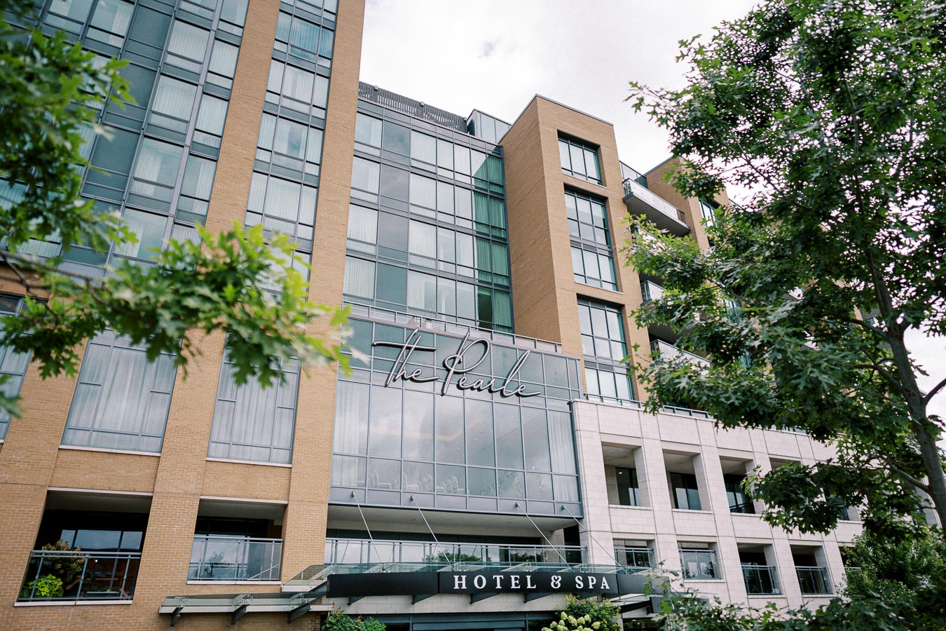 Modern hotel exterior with glass and brick facade surrounded by lush greenery, highlighting the inviting entrance to 'The Pearle Hotel & Spa' on a sunny day.