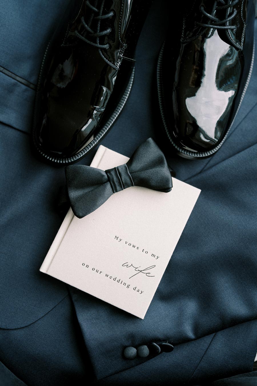 Black polished dress shoes, a silk bow tie, and a vow book titled My vows to my wife on our wedding day placed on a navy blue suit, representing elegant wedding attire for the groom.