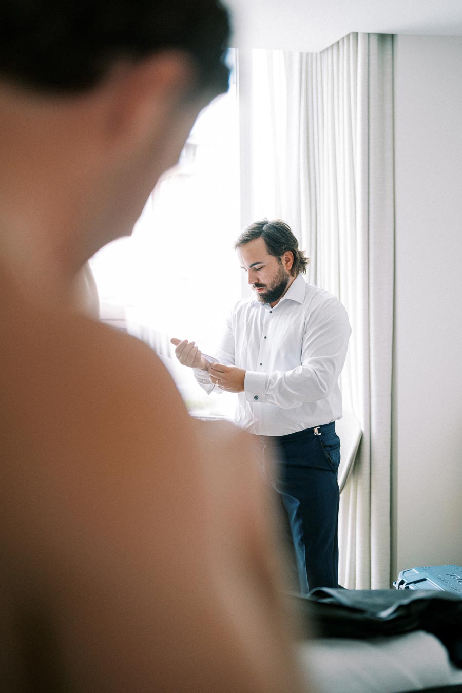Groom preparing for wedding, adjusting cufflinks in bright room with curtains.