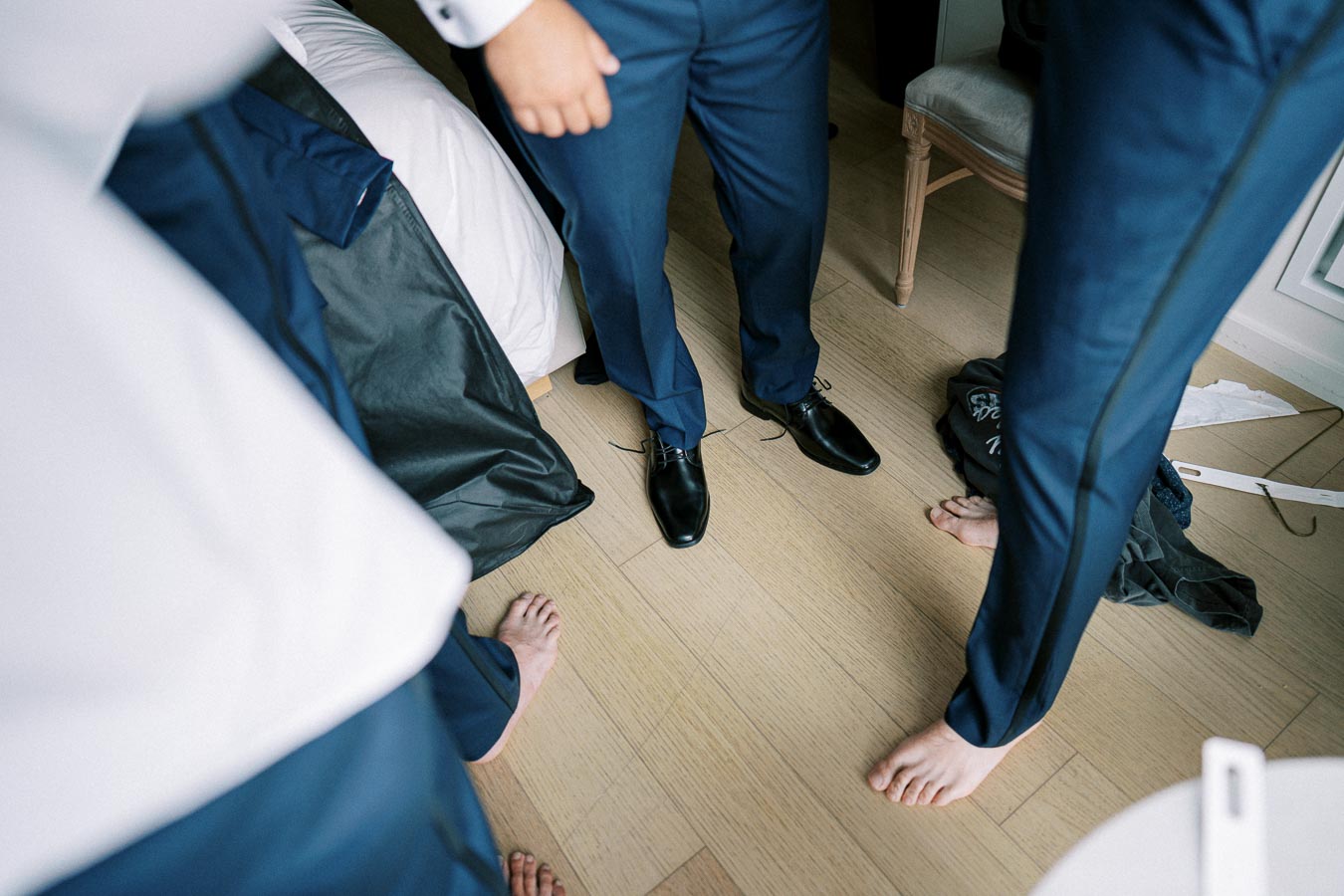 Formal menswear fitting in a hotel room, featuring individuals adjusting their navy blue suits and polished black shoes, highlighting the preparation process for a special event.