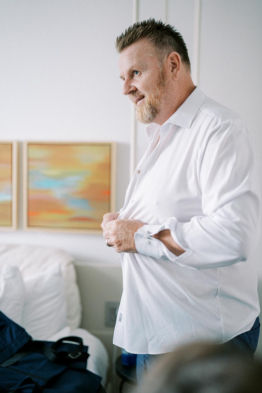 Man buttoning white shirt in modern bedroom setting, with abstract art on wall.
