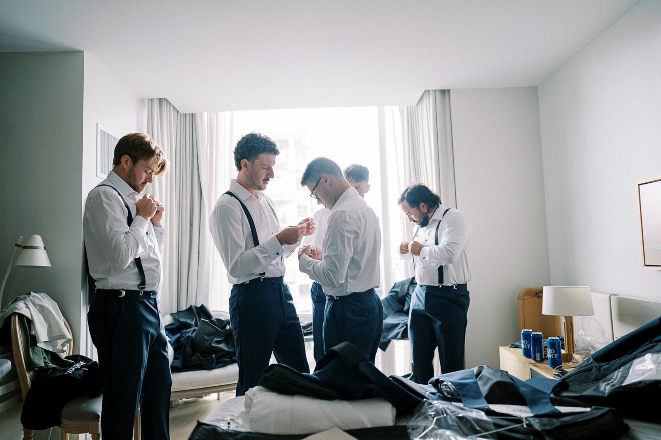 Group of groomsmen in a hotel room getting ready for a wedding, wearing white shirts and suspenders, with suits and accessories spread out on furniture.