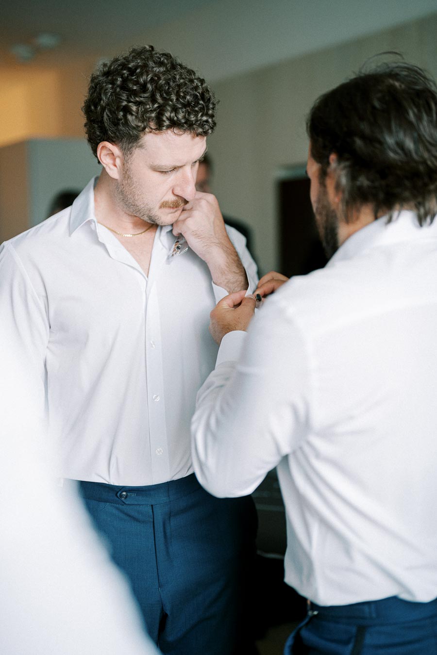 Groom getting ready with help from a groomsman, dressed in a white shirt and blue trousers, adjusting cufflinks before a wedding ceremony.